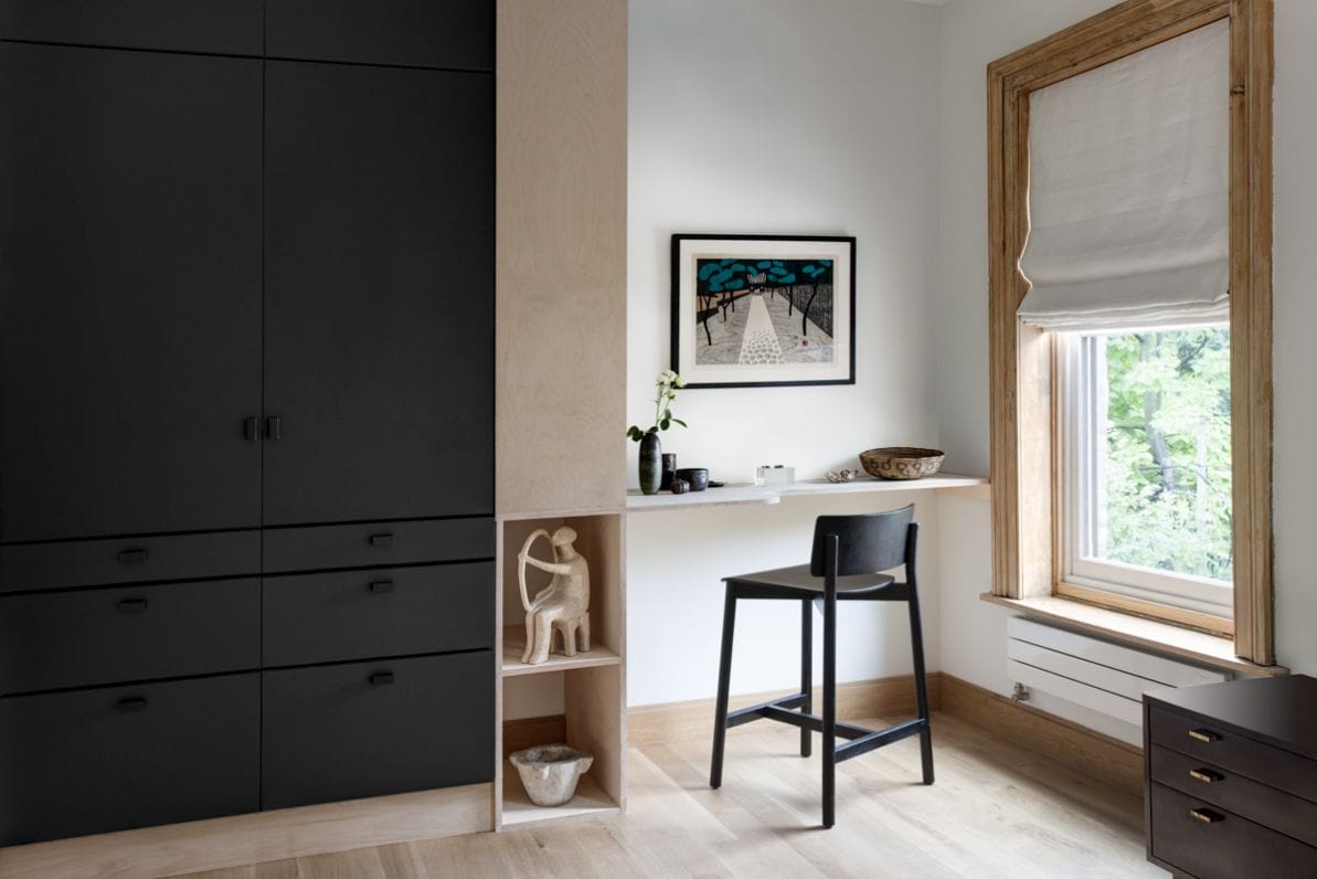 Minimalist home office nook with black built-in cabinets, a slim white desk, black stool and wood-trimmed window